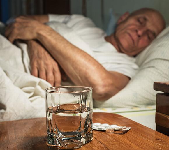 Elderly man sleeping with glass of water and tablets on table in front of him